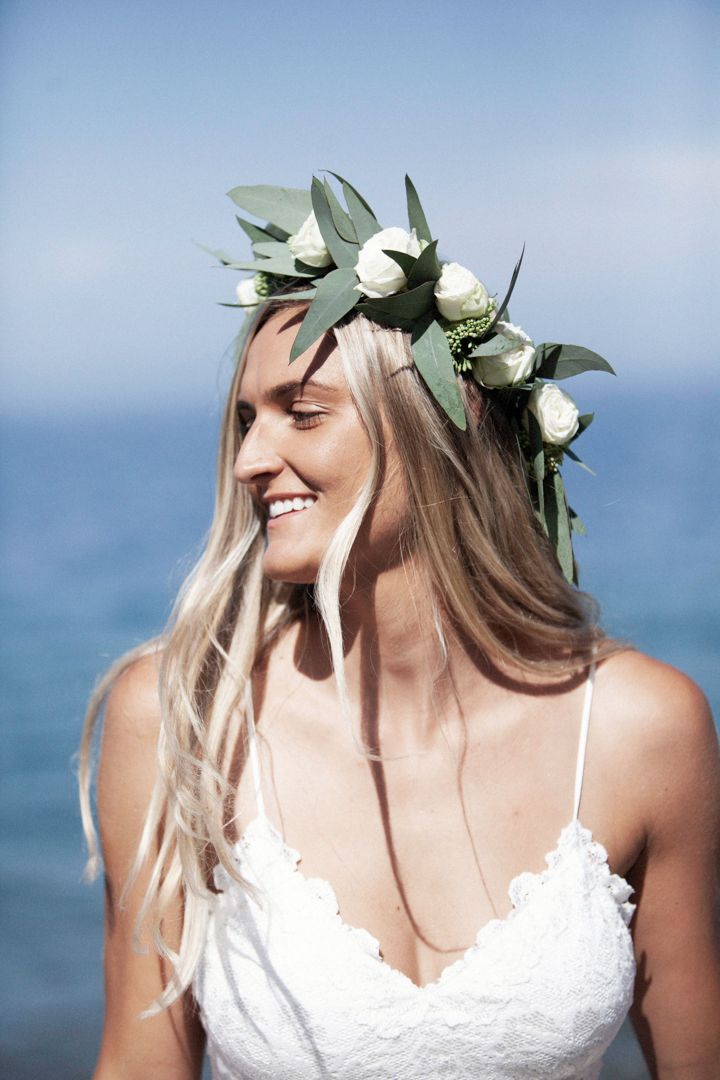 A bride wearing a delicate flower crown and white lace dress smiles against an ocean backdrop.