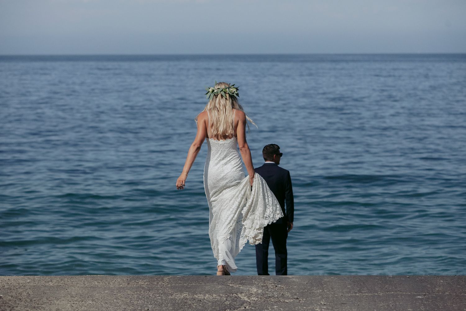 A romantic couple in wedding attire walk together along the shoreline with turquoise ocean waters in the background.