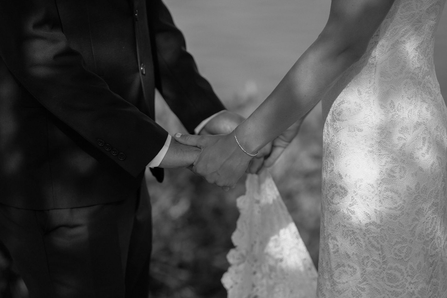 Black and white close-up of newlyweds holding hands, showing a bride in lace and groom in dark formal attire.