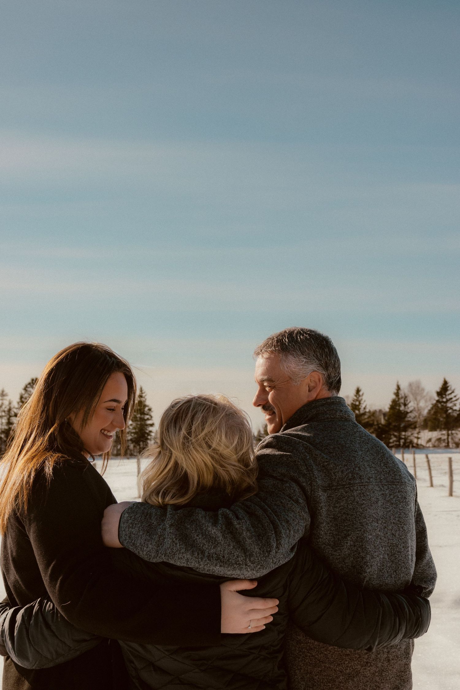 Three people share an intimate embrace while standing in a snowy field with pine trees in the background.