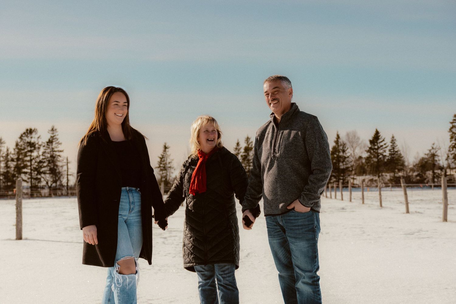 Three people hold hands while standing together in a snowy field with evergreen trees.