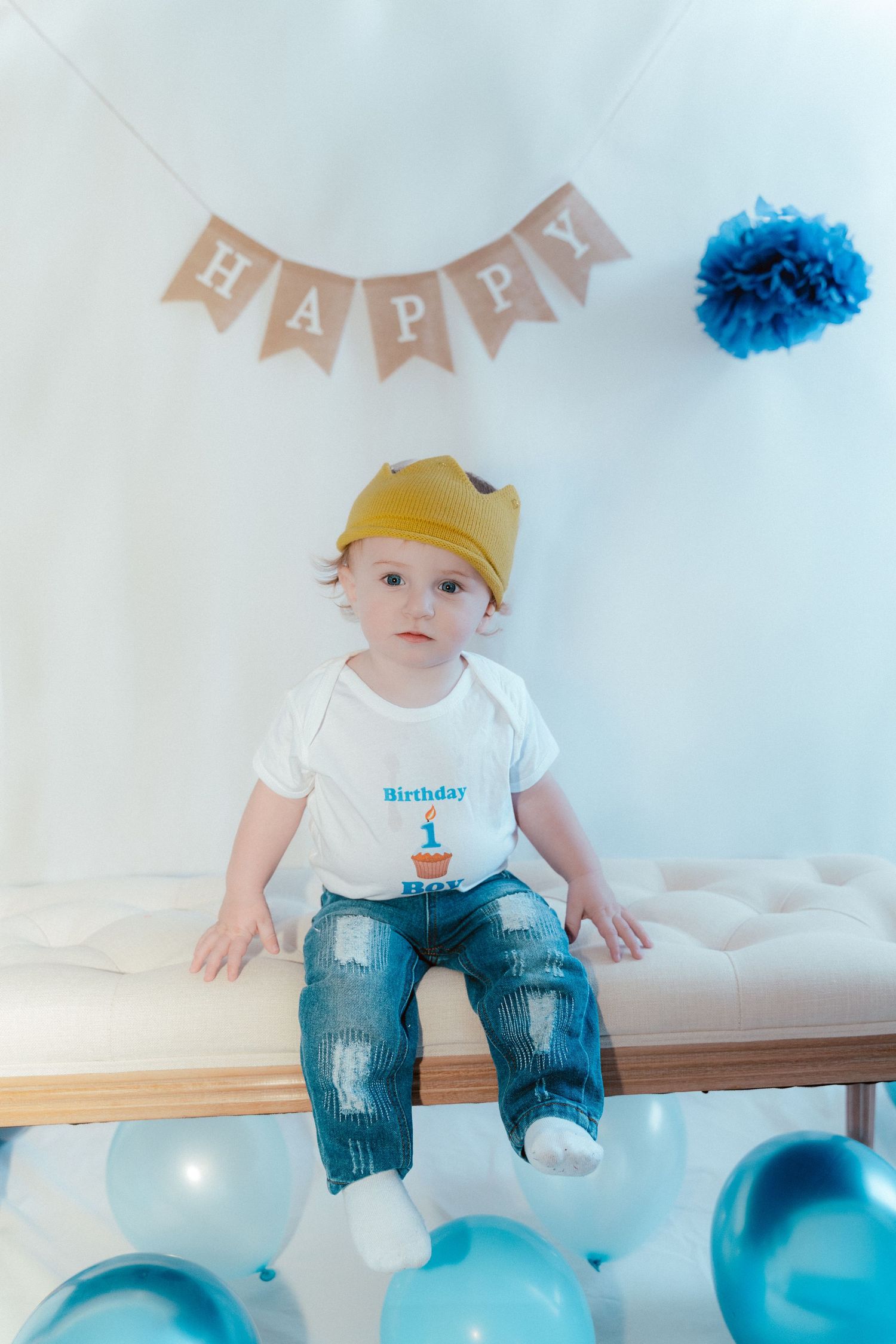 Baby in white shirt and blue jeans sits with turquoise party balloons and gold crown for first birthday celebration.