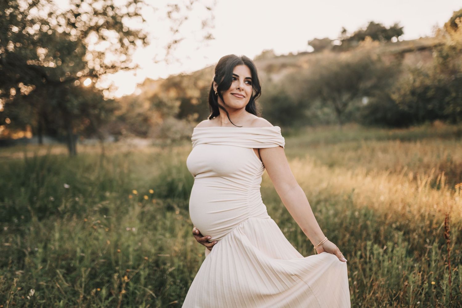 Someone poses in a flowing off-shoulder cream dress during a golden hour maternity photoshoot in a grassy field.