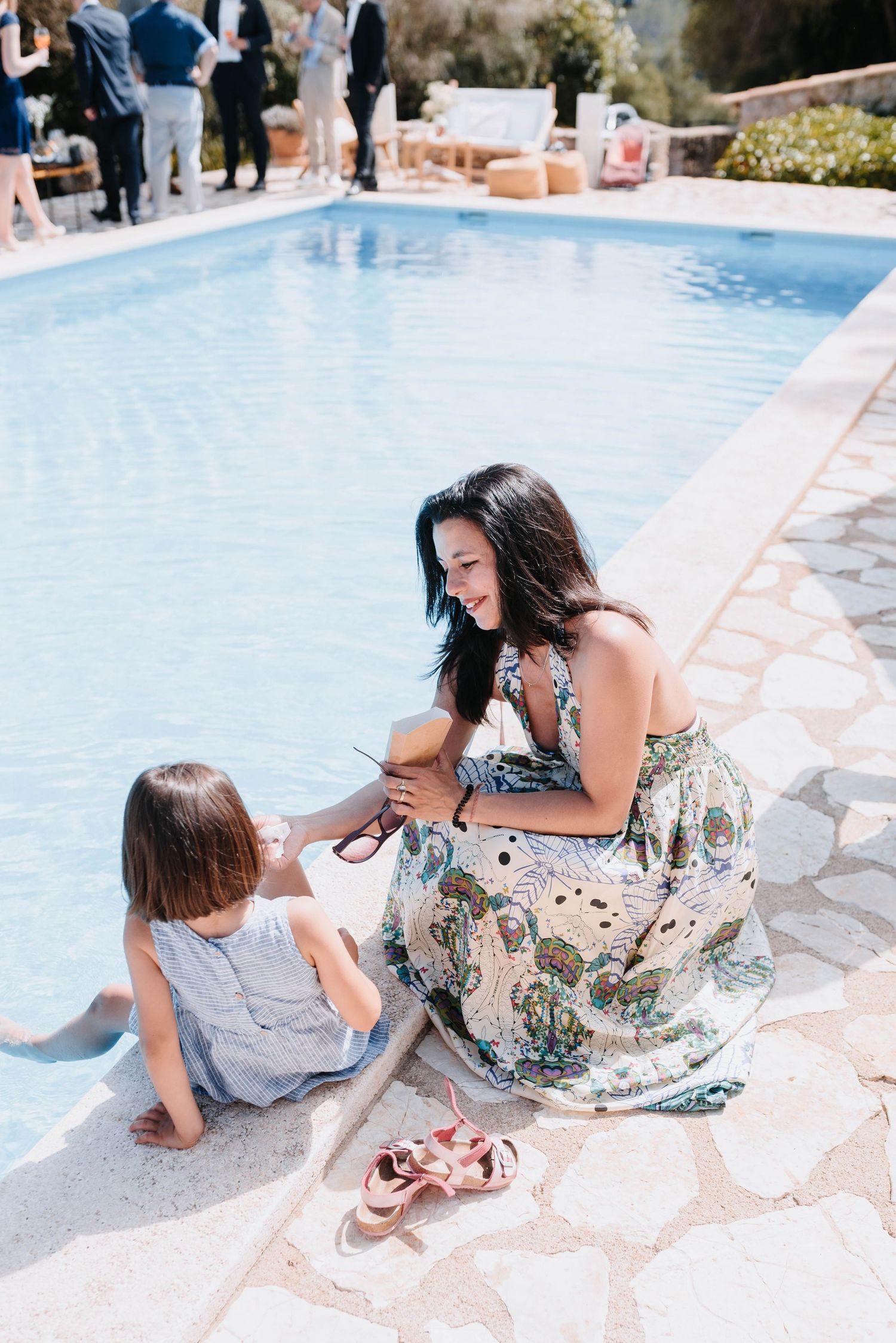 Eine Mutter und ihre Tochter genießen einen sonnigen Tag am Pool, während das Kind spielt und ihr Blumenkleid im Wind weht.