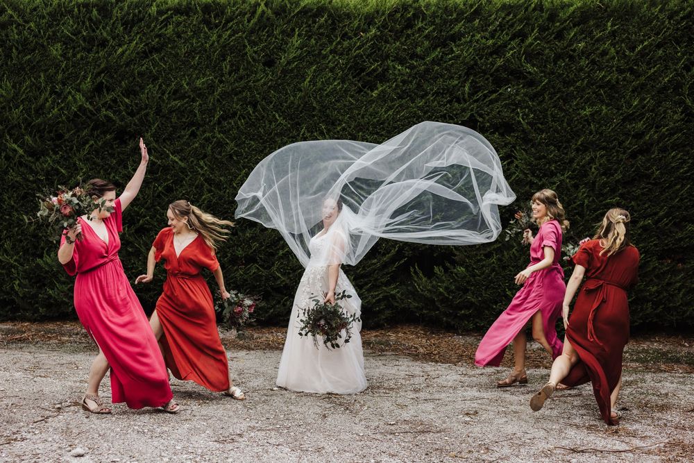 Wedding party in red dresses react playfully as wind catches bride's veil during outdoor photo session.