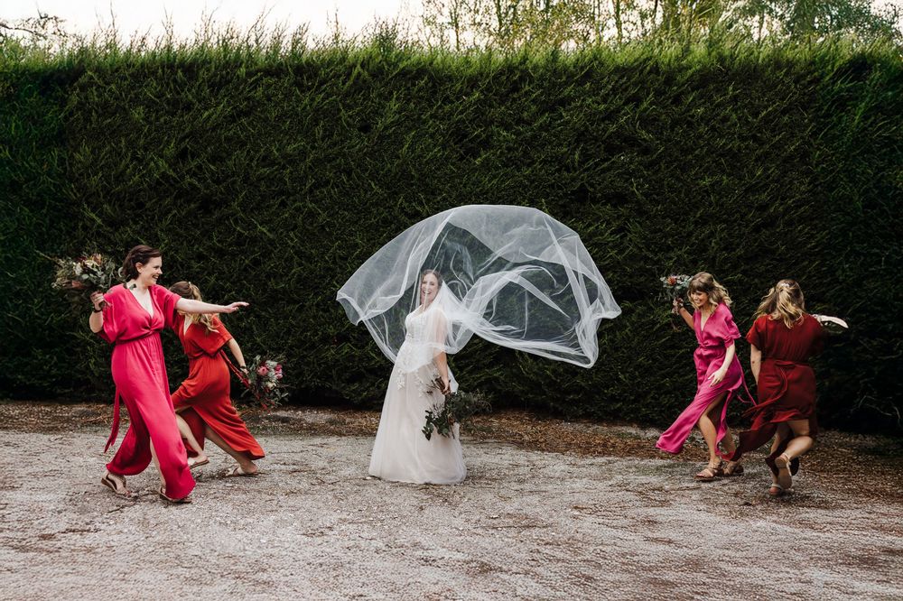 Wedding party dances joyfully outdoors as veil flows in the wind against green hedges.