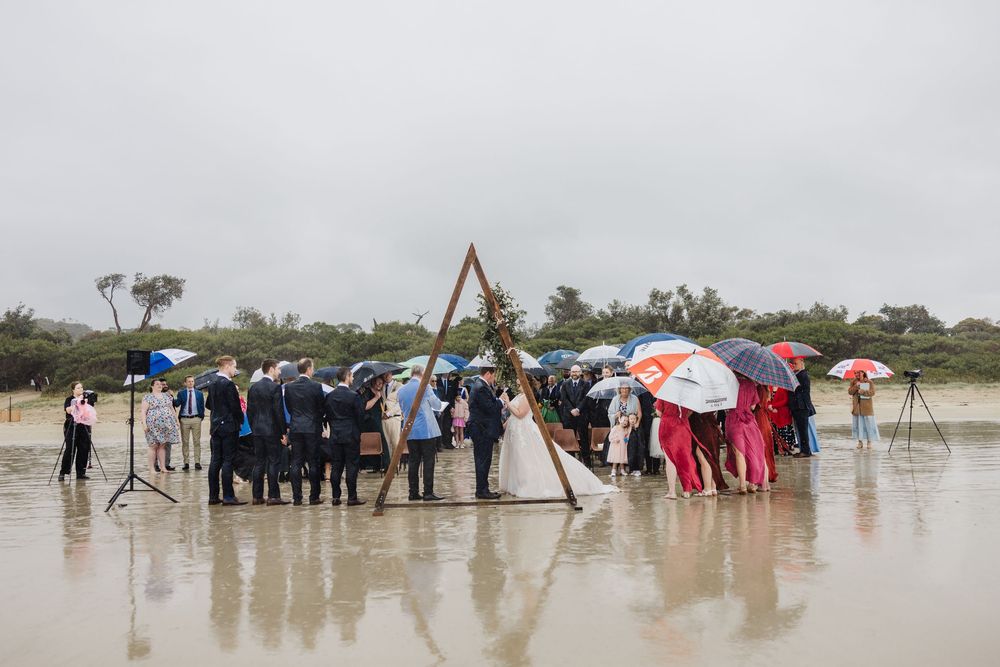 Wedding ceremony on rainy beach with guests holding umbrellas under wooden arch structure.