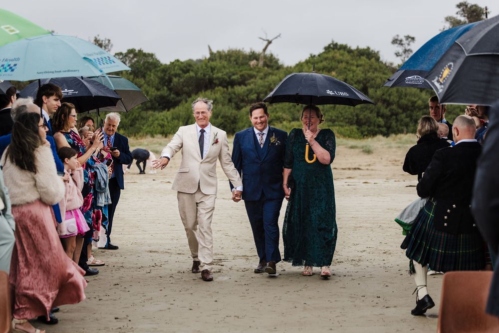 Wedding guests holding umbrellas walk on a sandy beach path during an outdoor ceremony in misty weather.