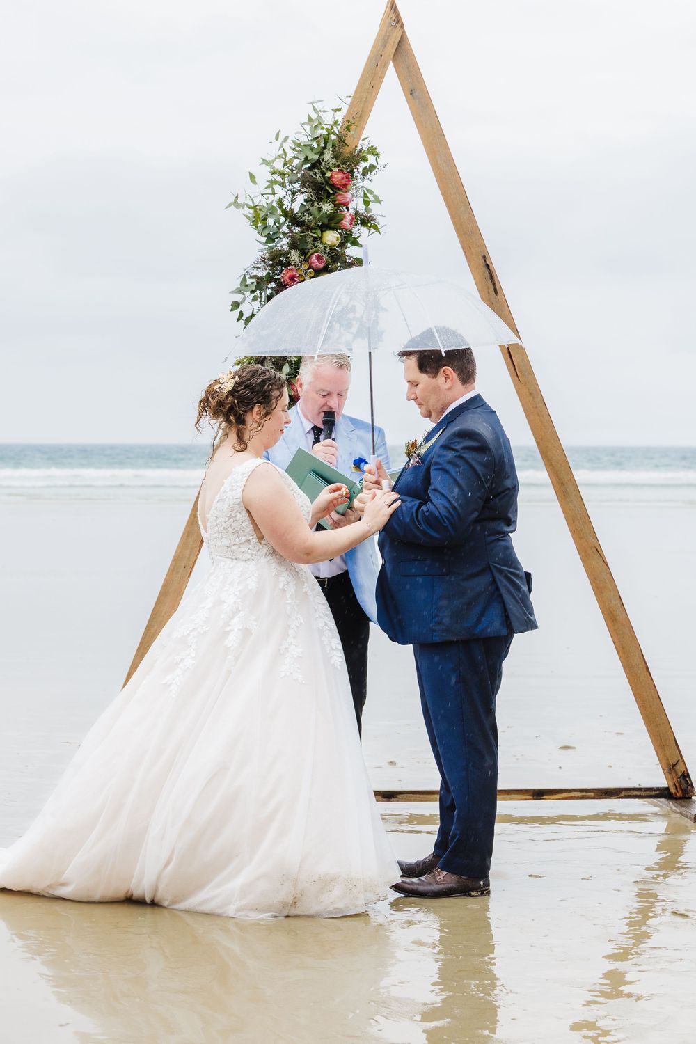 A wedding ceremony takes place on a beach under a wooden triangle arch with a white umbrella and floral decor.