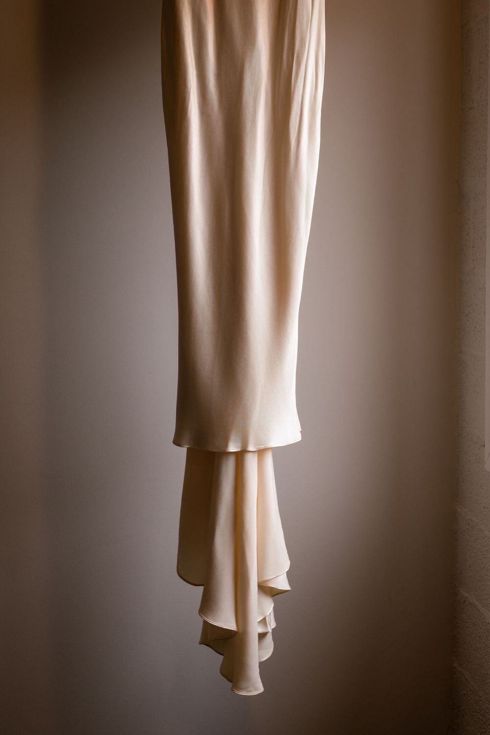 Elegant cream satin wedding dress hem hanging, showcasing delicate draped fabric layers in soft warm lighting.