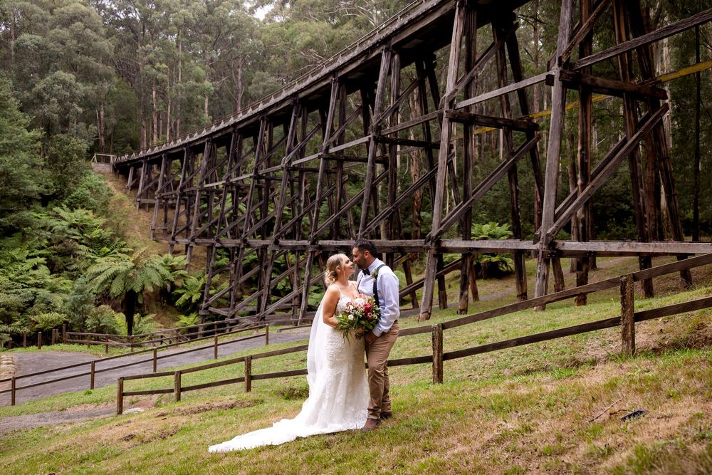 Bride and groom kissing beneath a historic wooden trestle bridge surrounded by lush green forest.