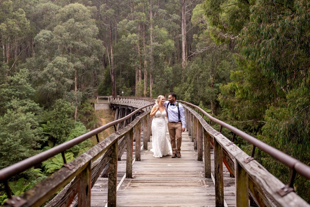 Couple walking hand in hand on a wooden bridge surrounded by lush green forest on their wedding day.