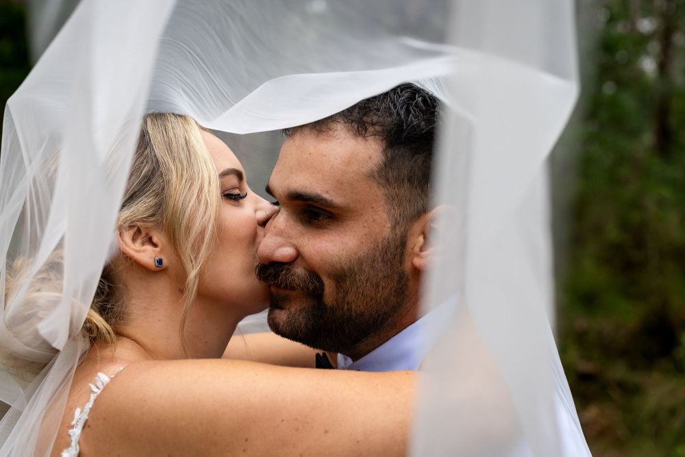 Bride and groom sharing a romantic kiss under a flowing white wedding veil outdoors.