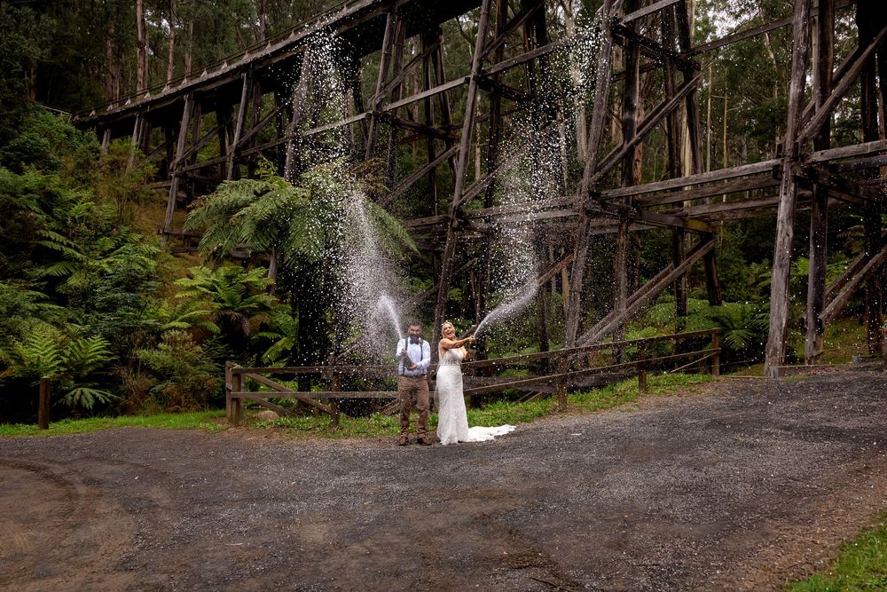 Bride and groom celebrate with champagne spray near a historic wooden trestle bridge surrounded by lush forest ferns.