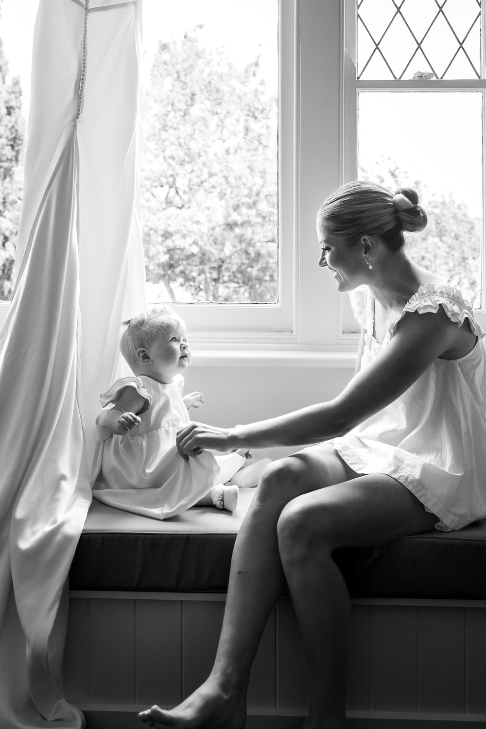 Black and white photo of a young mother sitting on a window seat, smiling and holding hands with her baby girl.