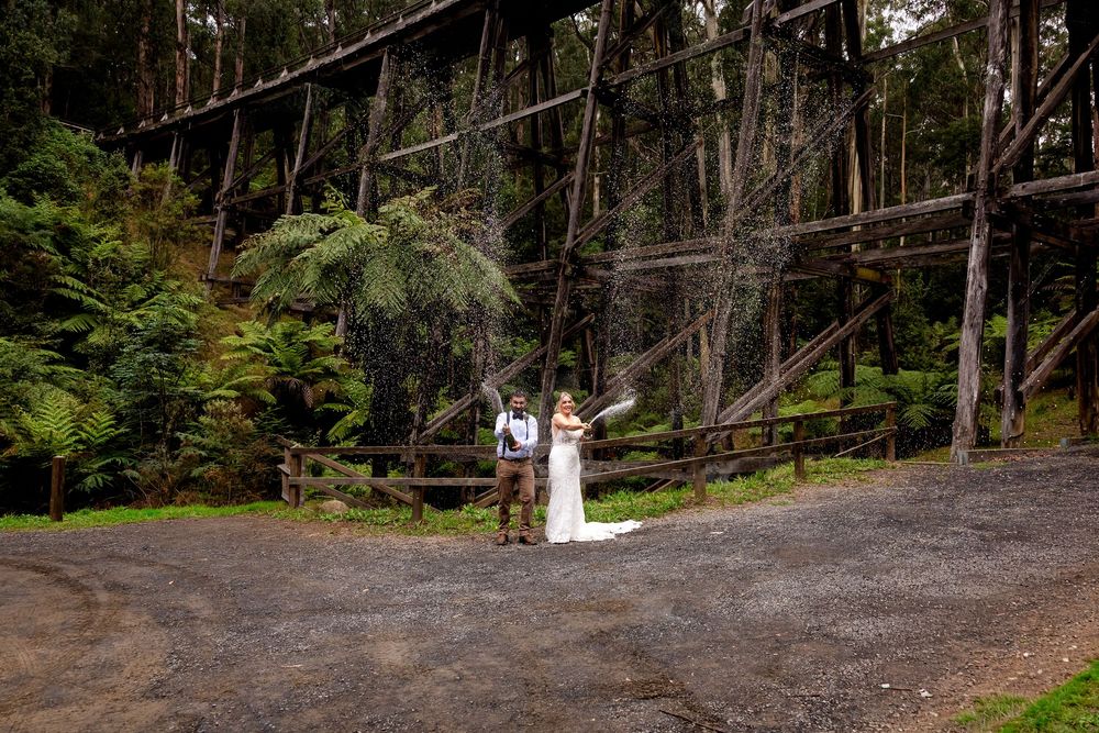 Bride and groom stand together in a lush forest setting beneath a historic wooden trestle bridge.