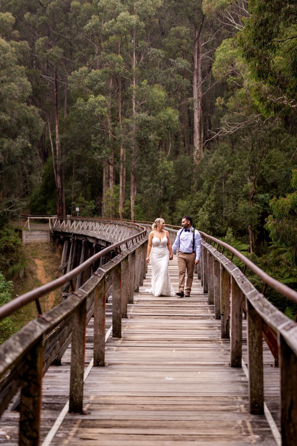 Bride and groom walking hand in hand on a rustic wooden bridge surrounded by tall trees in a lush forest setting.