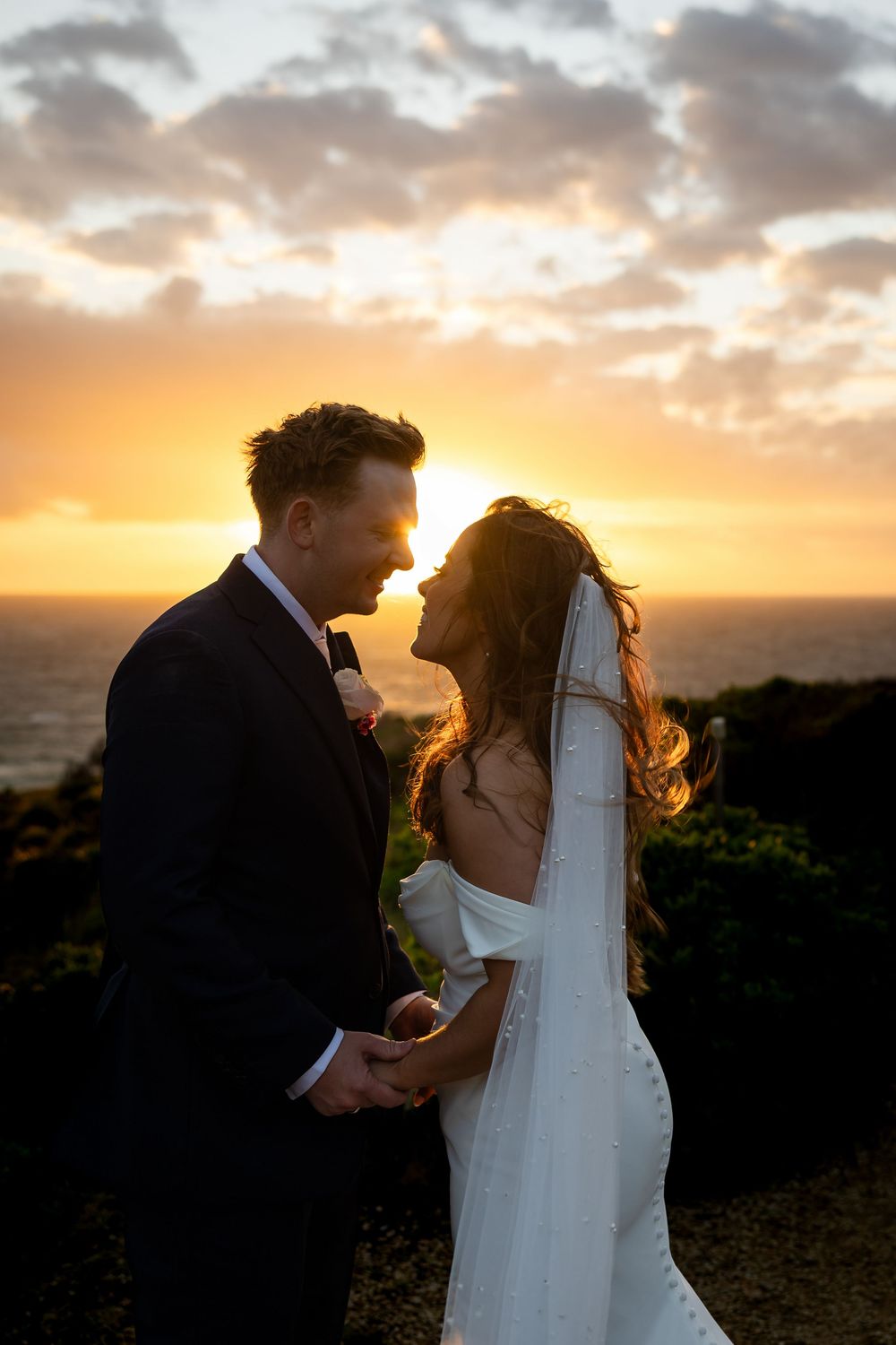 Bride and groom share a romantic moment at sunset with ocean views, golden light glowing behind them on their wedding day.