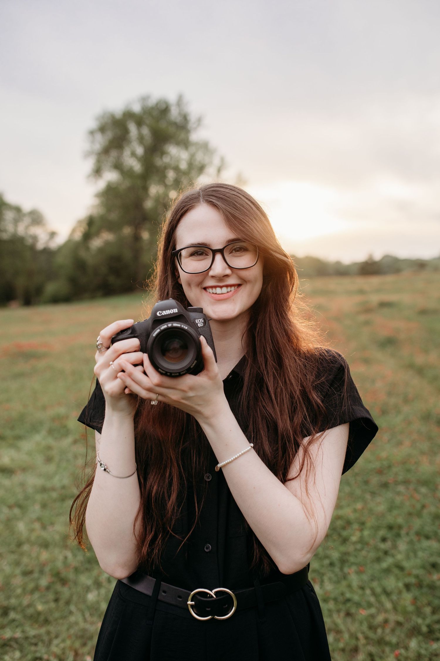 @oliviapaigephoto Olivia from Olivia Paige Photography holding a camera and smiling in a branding photo taken in a field at sunset
