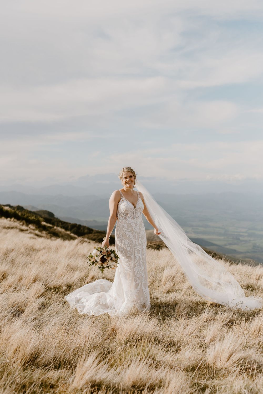A bride in a lace wedding gown with flowing veil stands on a windswept hilltop holding a bouquet against misty mountains.