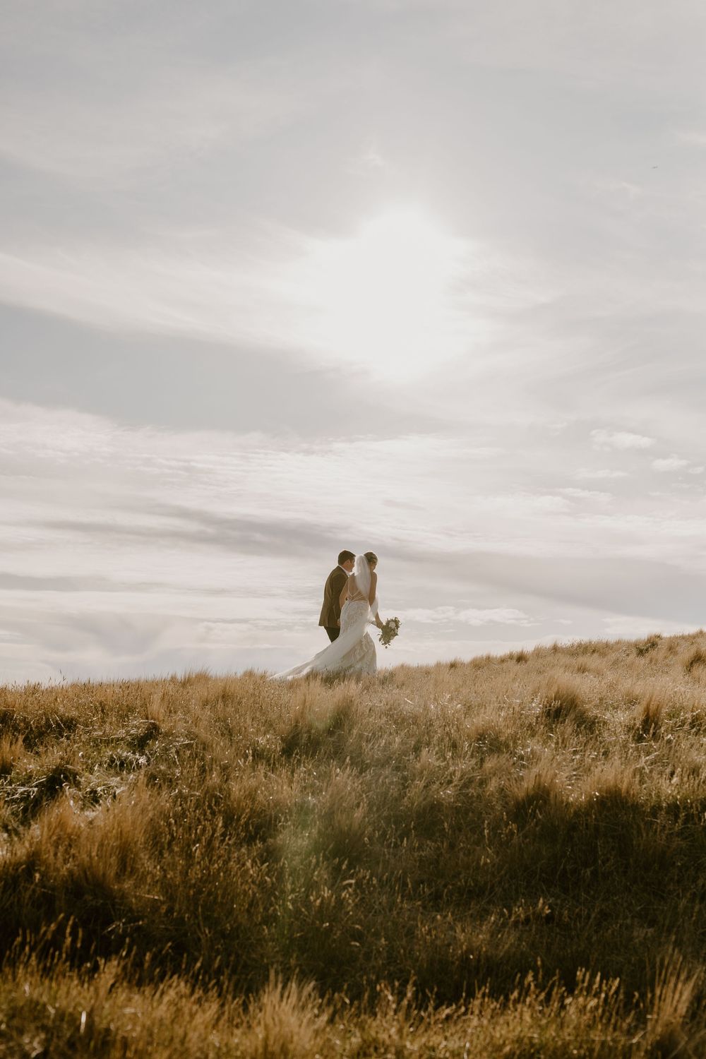 A couple stands together on a hillside silhouetted against a soft cloudy sky during golden hour.