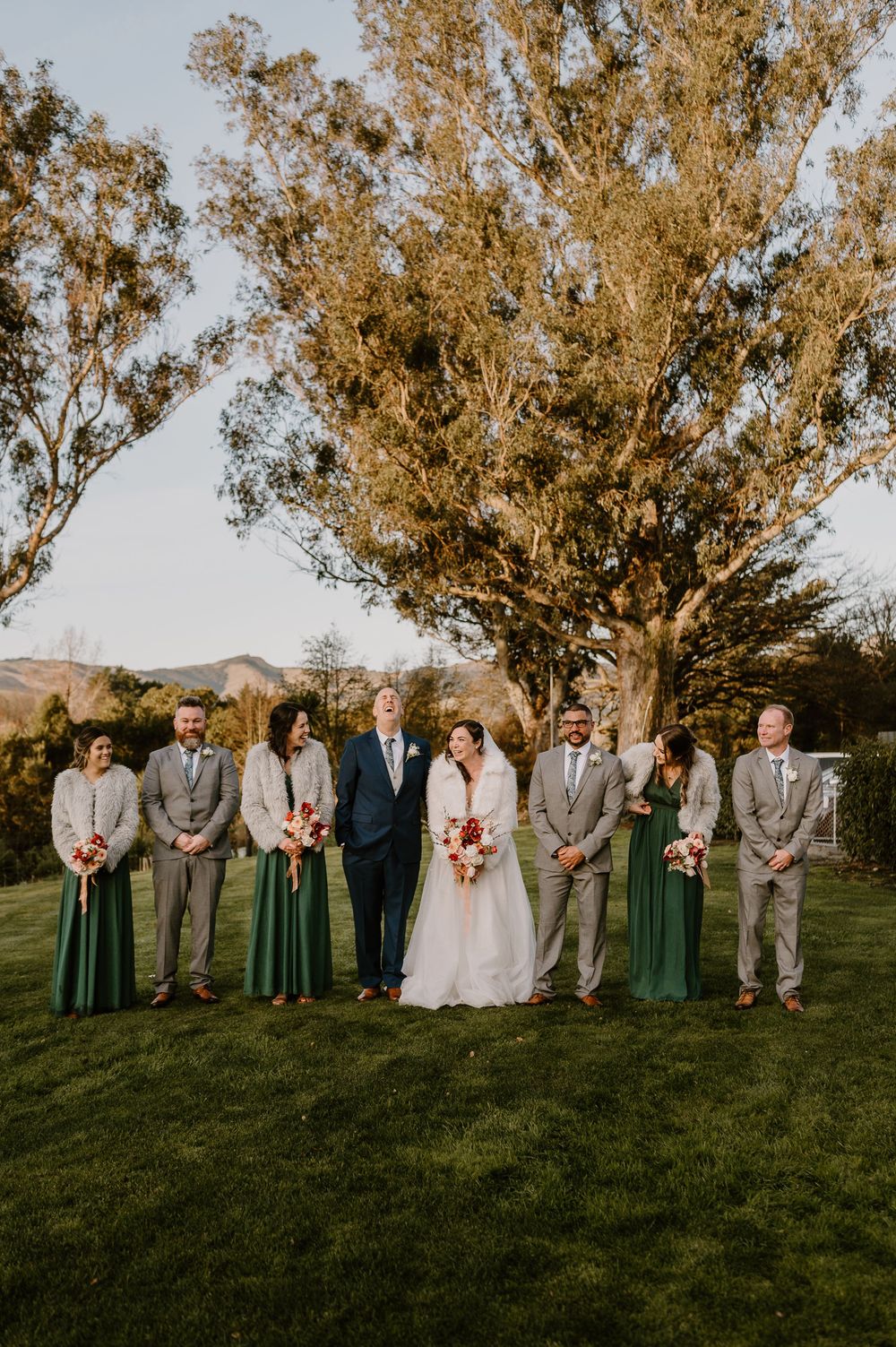 Wedding party stands in front of large tree at sunset wearing sage green and grey attire for outdoor ceremony.