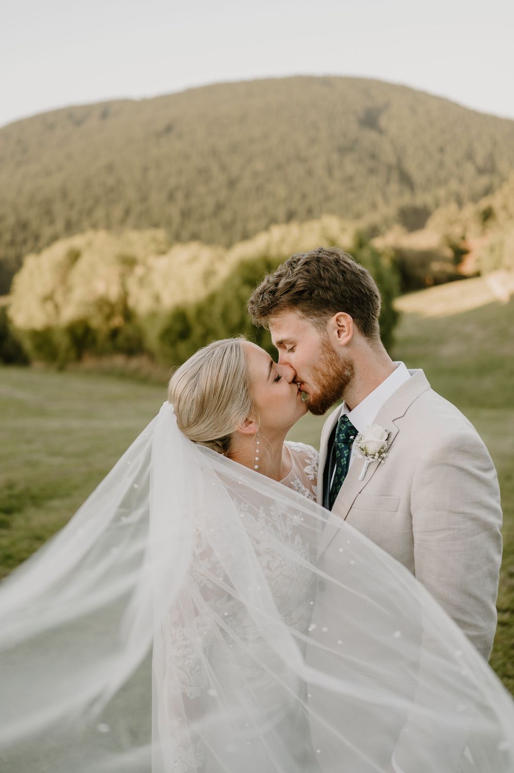 A romantic wedding couple shares a kiss under a flowing veil with mountains and greenery in the background.