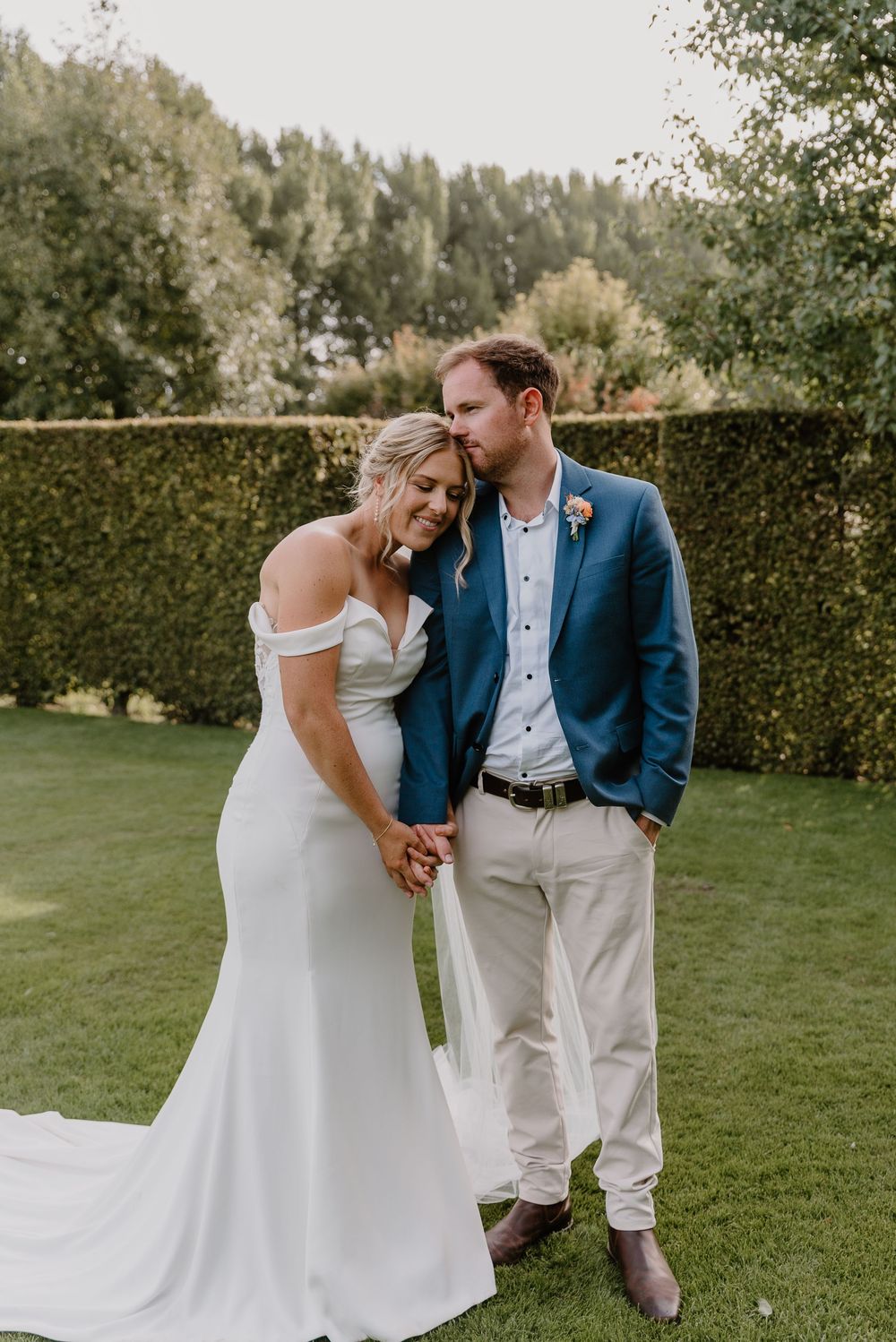A couple shares a sweet moment on a green lawn surrounded by lush hedges and trees during an outdoor wedding celebration.
