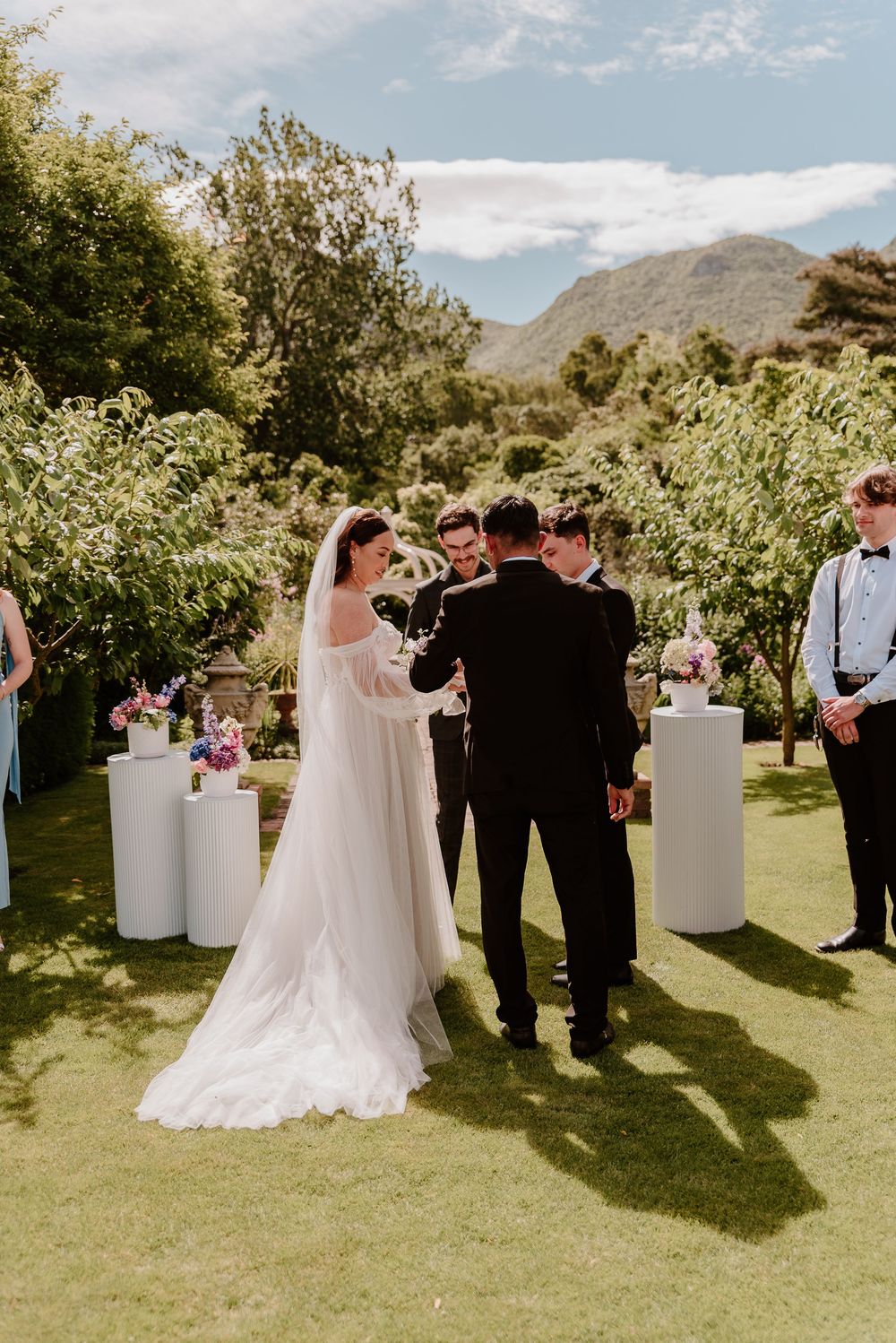 Wedding ceremony at an outdoor vineyard venue with white pedestals arranged on a green lawn against mountain backdrop.