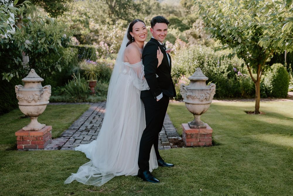 A couple in wedding attire poses in a garden setting with decorative stone urns on brick pedestals.