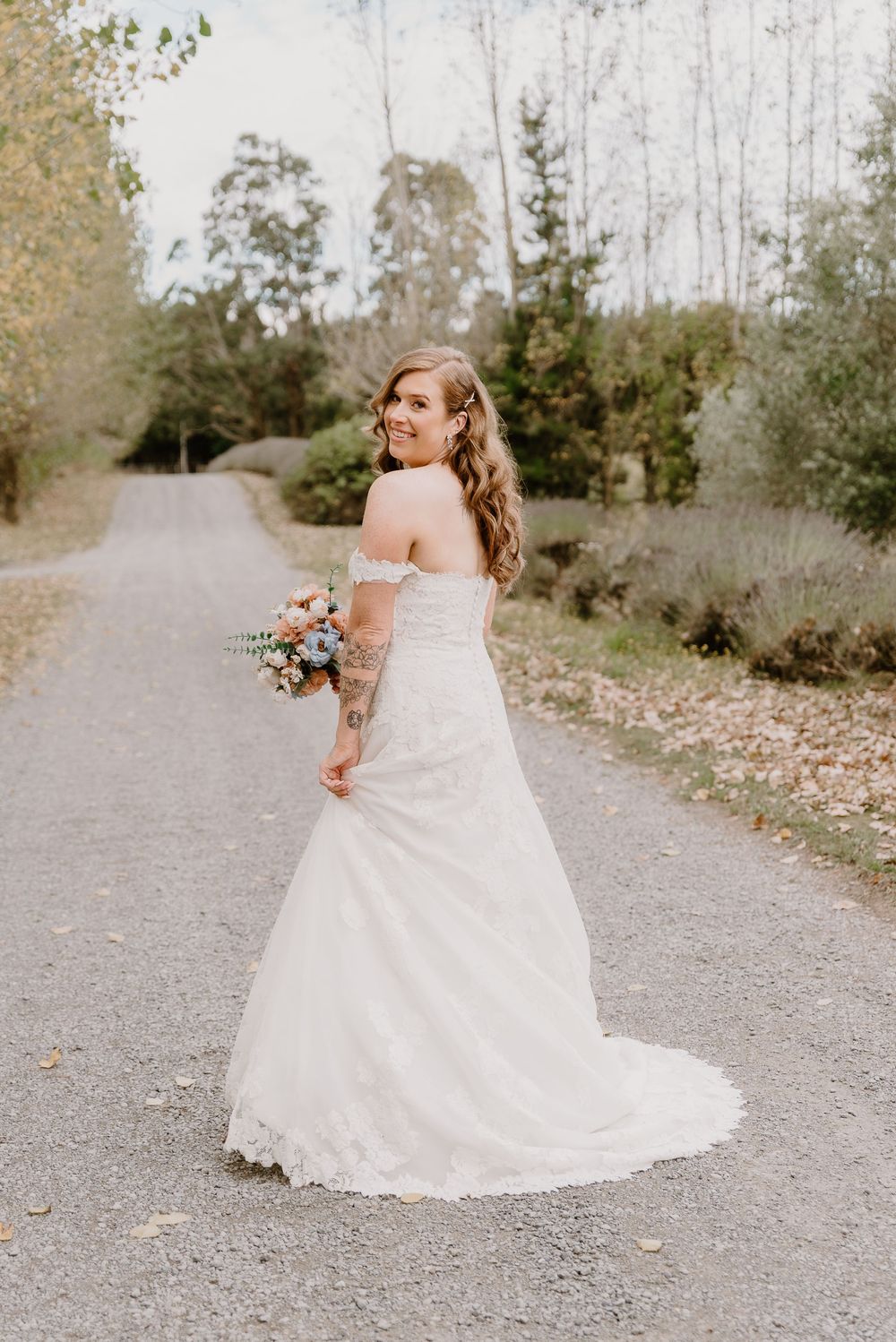 A bride in a stunning white lace strapless gown holds a bouquet while standing on a gravel path surrounded by trees.