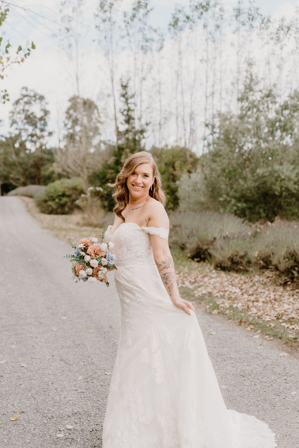 A bride in a beautiful off-shoulder lace wedding gown holds a romantic peach and white bouquet on a country path.