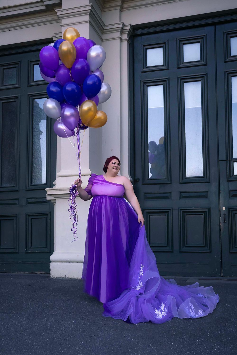 Elegant purple formal gown with flowing train paired with coordinating purple, gold and silver balloons against dark doors.