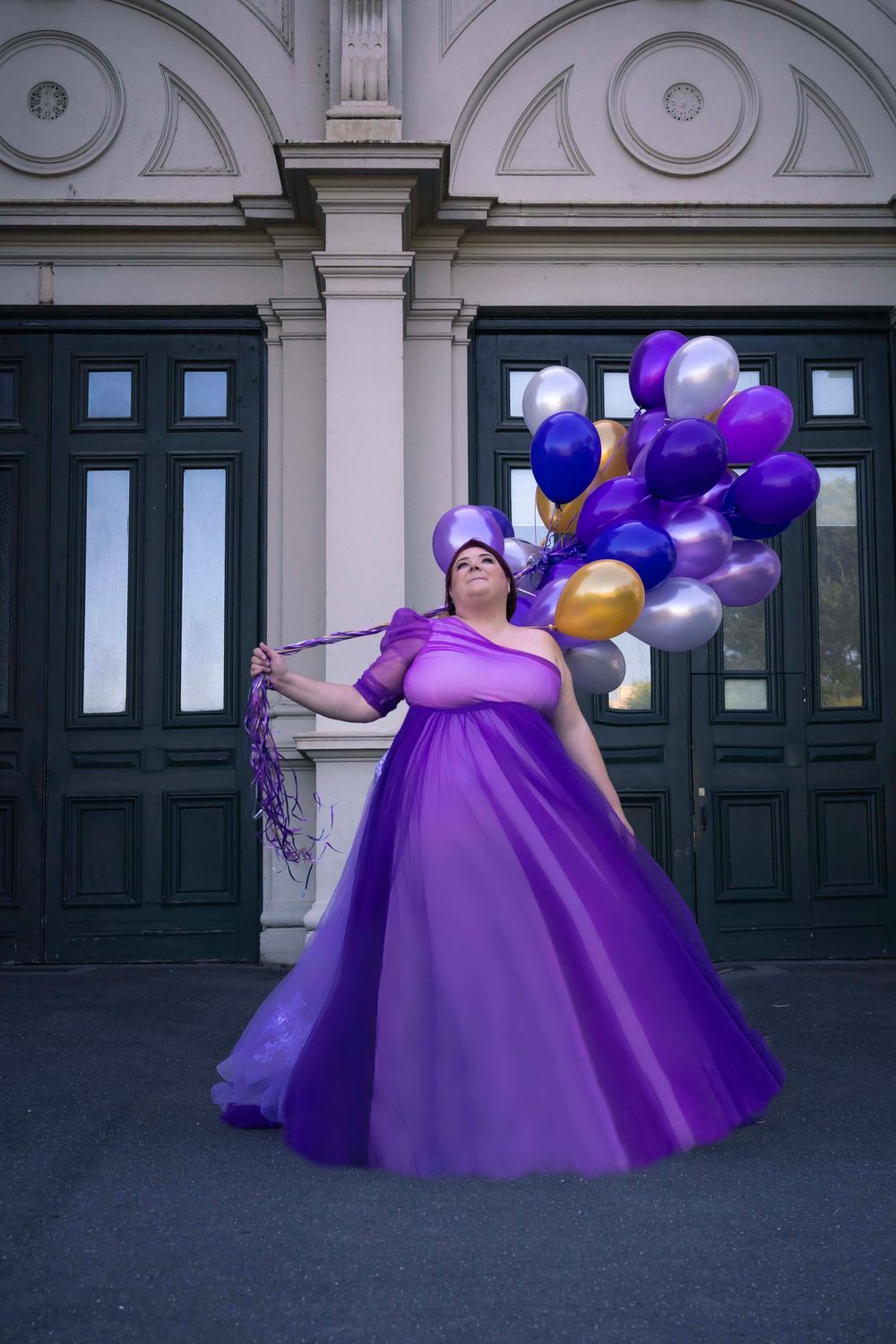 A figure in a flowing purple gown poses with purple and gold balloons against a grand doorway with classical architecture.