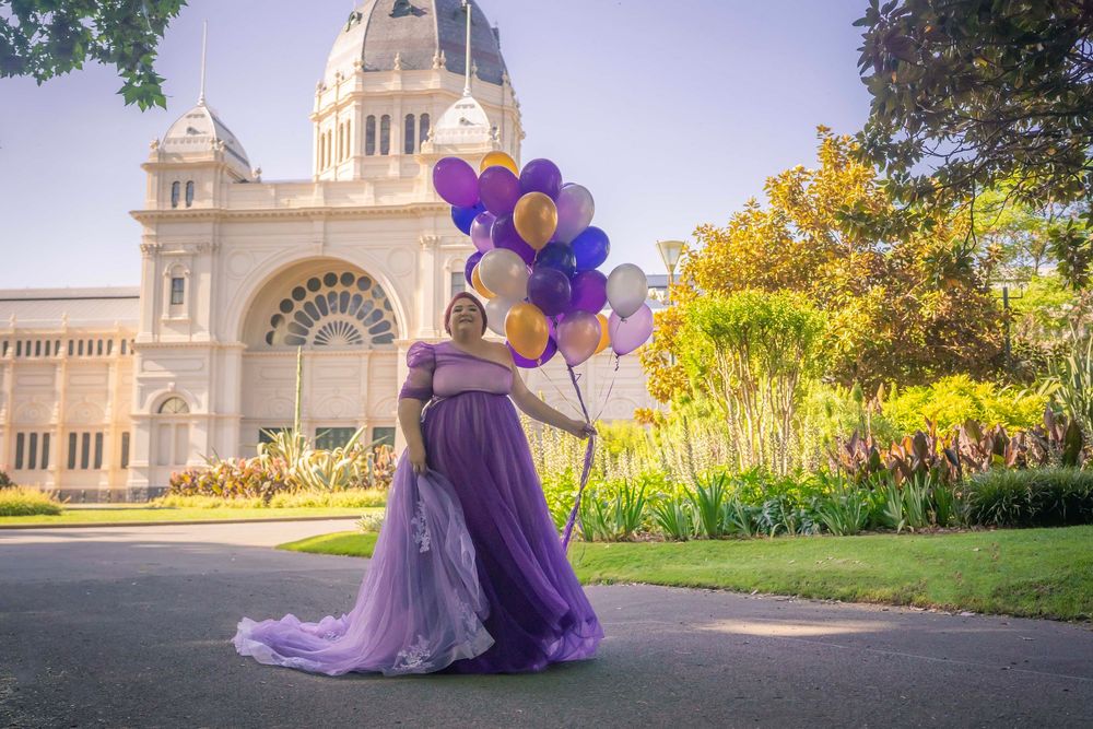 A figure in a flowing purple dress holds purple and gold balloons in front of a grand historic building with a domed tower.