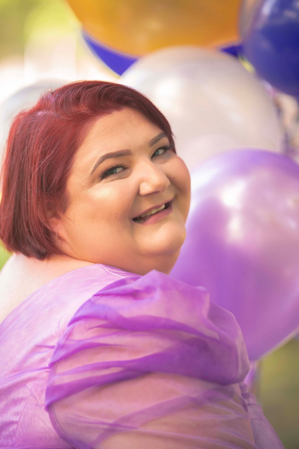 A joyful person with vibrant red hair poses with colorful balloons while wearing a pink flowing dress.