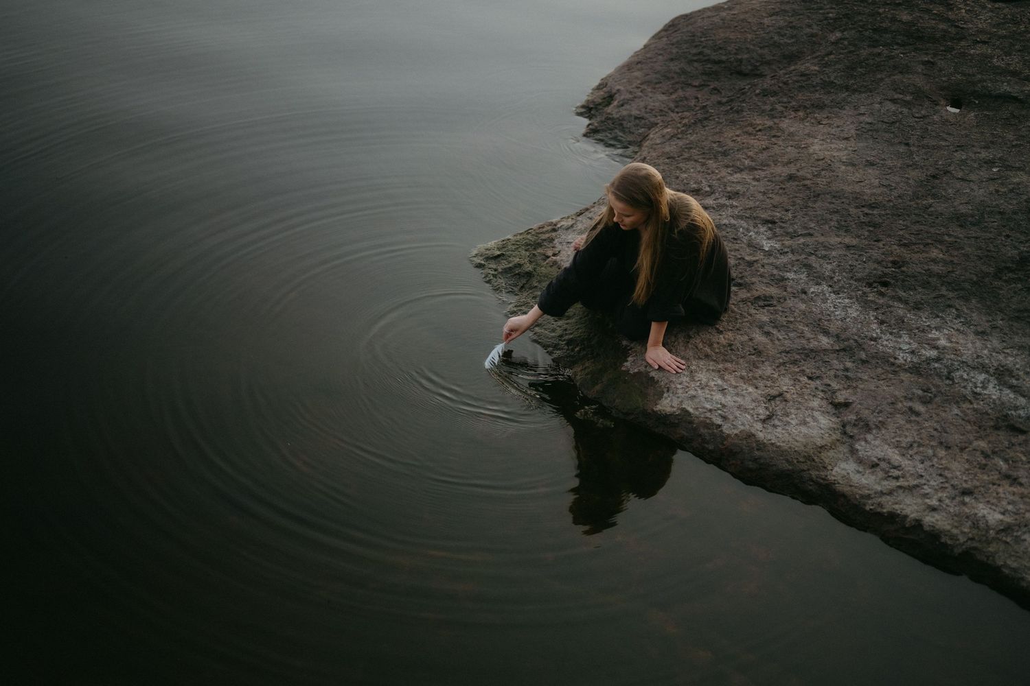 A dog swims in a dark lake while splashing water near a rocky shoreline on a cloudy day.