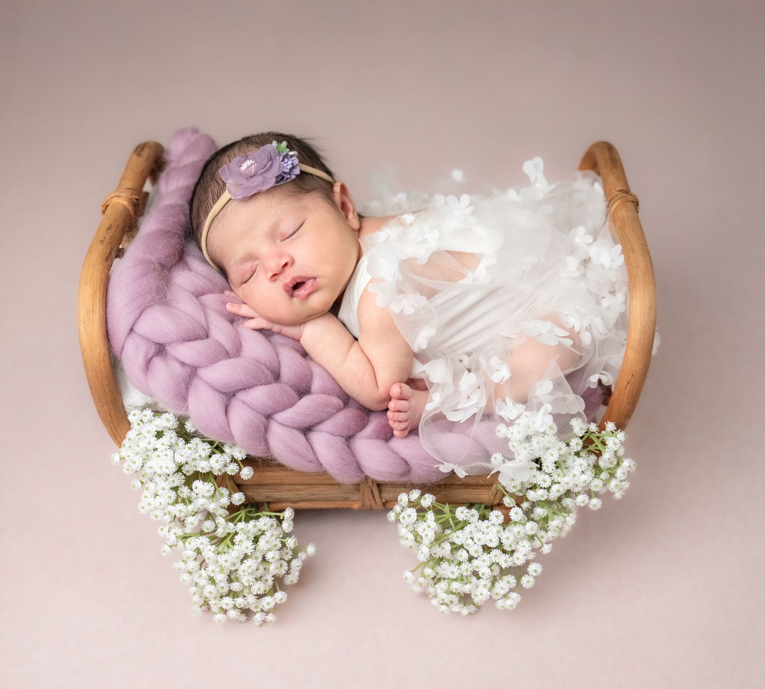A sleeping baby nestled in a wooden bowl with purple knit blanket and baby's breath flowers.