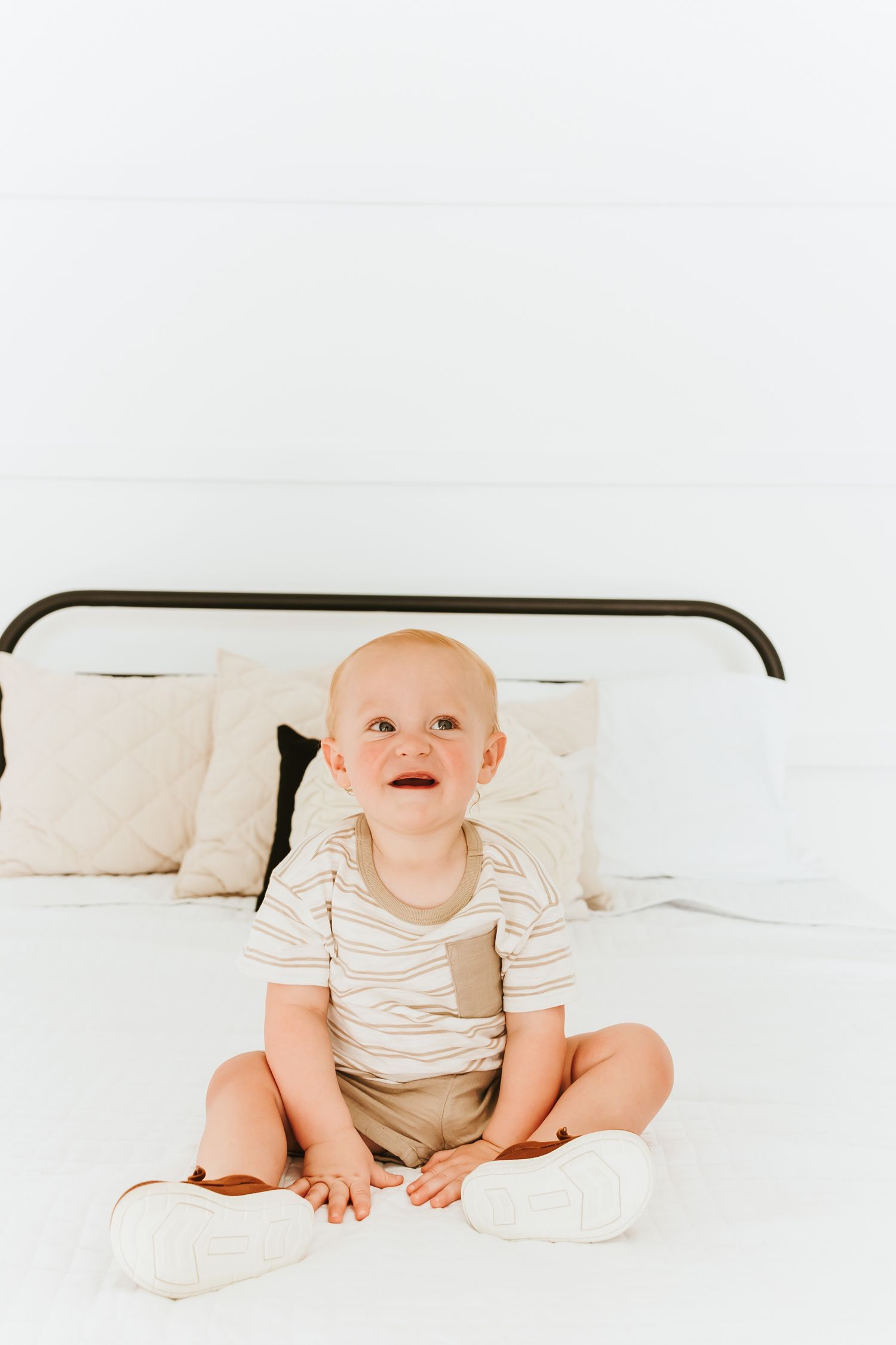 A happy baby in striped clothing sits on a white bed with pillows against a headboard.