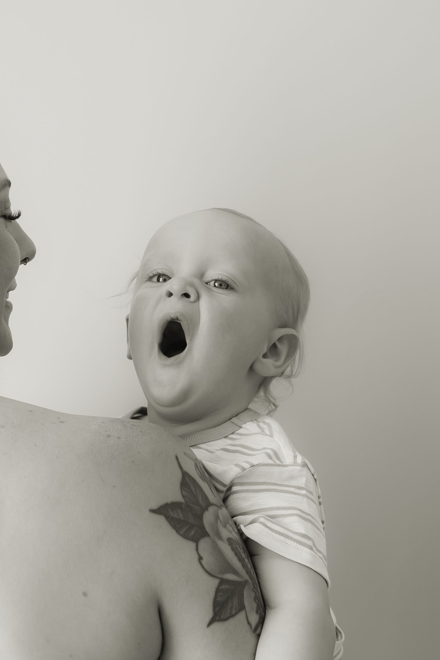 A black and white portrait showing a baby yawning while positioned next to someone with visible tattoo artwork.