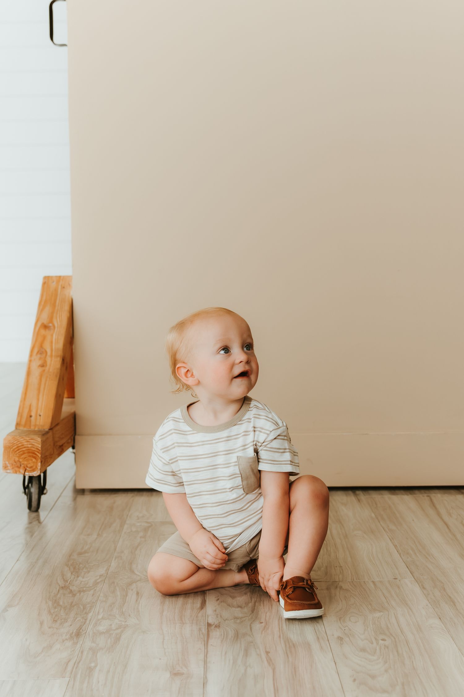 A baby in a striped onesie sits on light wooden flooring against a beige wall.