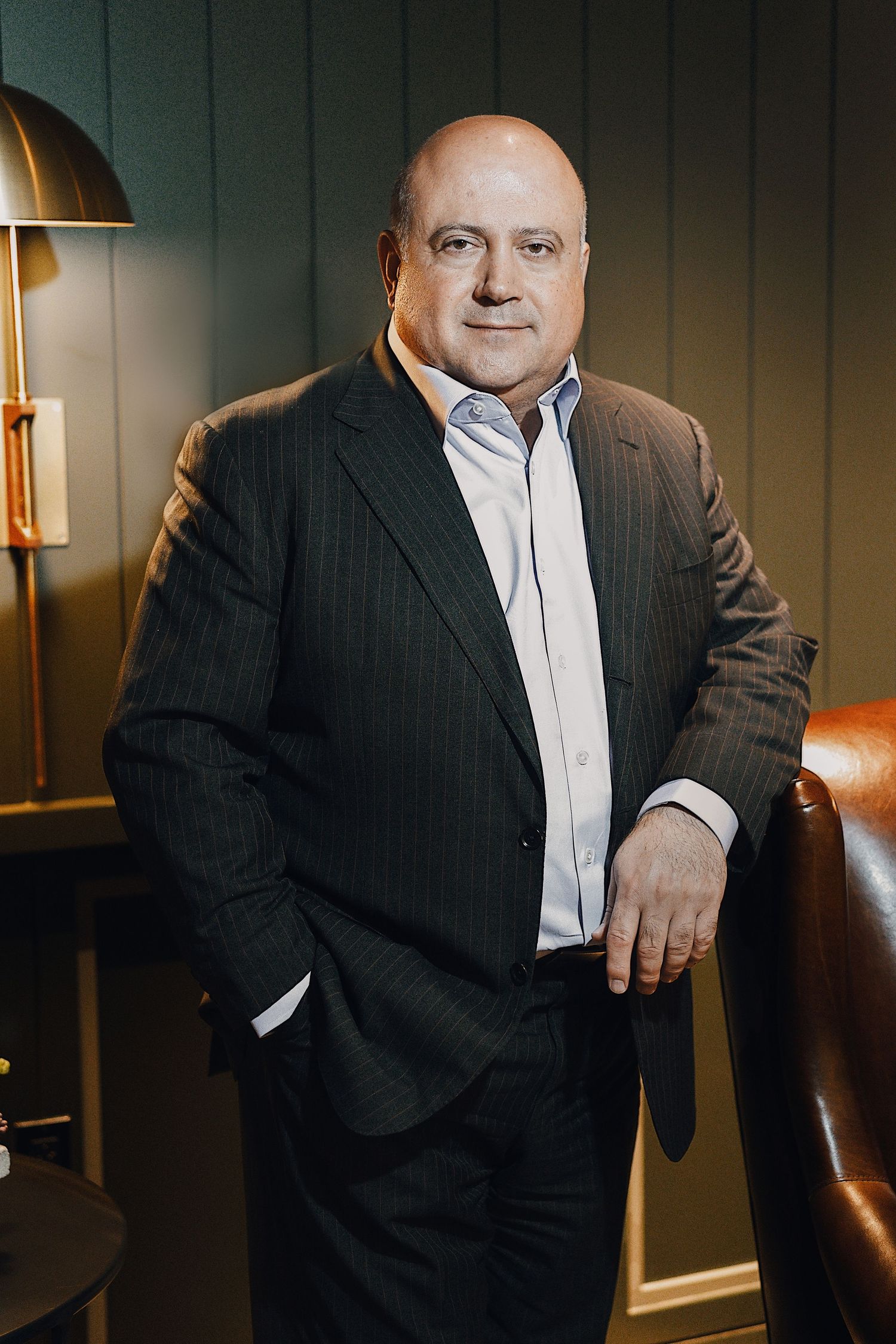 Bald businessman in dark pinstripe suit leaning on leather chair in moody setting.