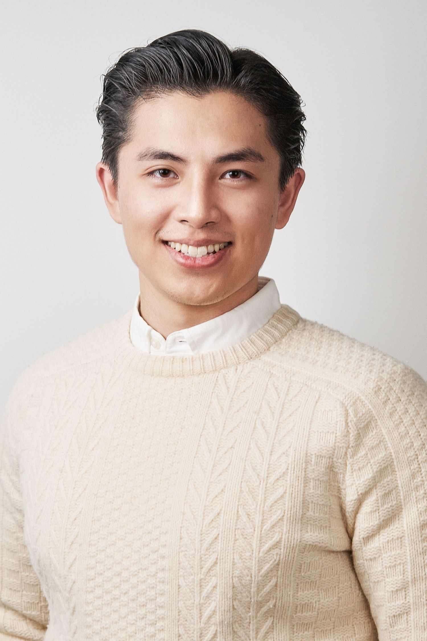Smiling young Asian man in cream cable-knit sweater poses for clean professional headshot.