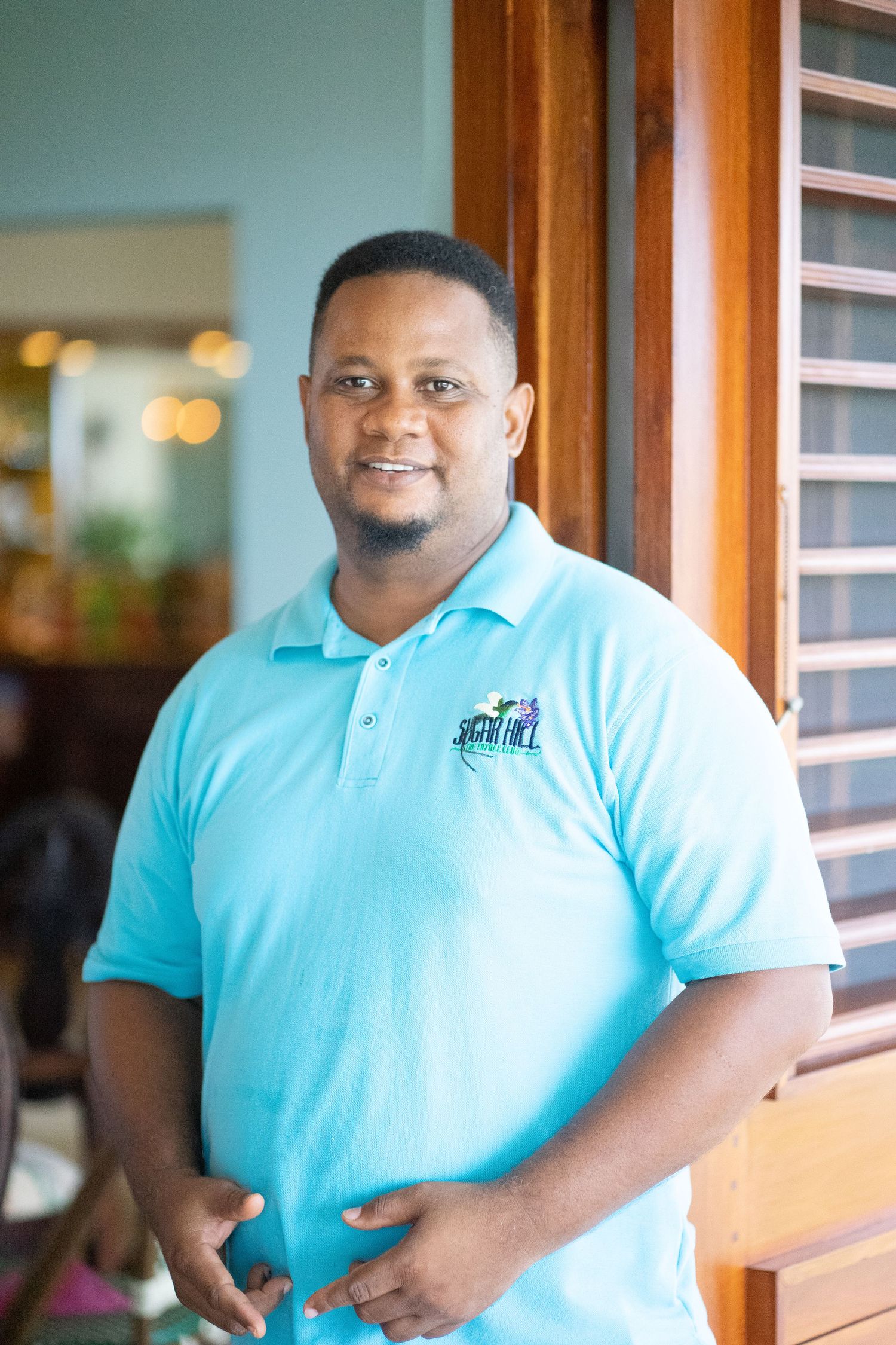 Smiling man in light blue polo shirt standing indoors near wooden shuttered windows in a bright setting.