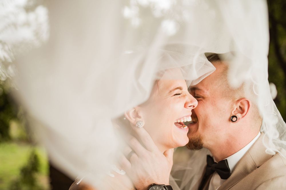 Joyful couple laughing together under flowing white veil during outdoor wedding photo session.