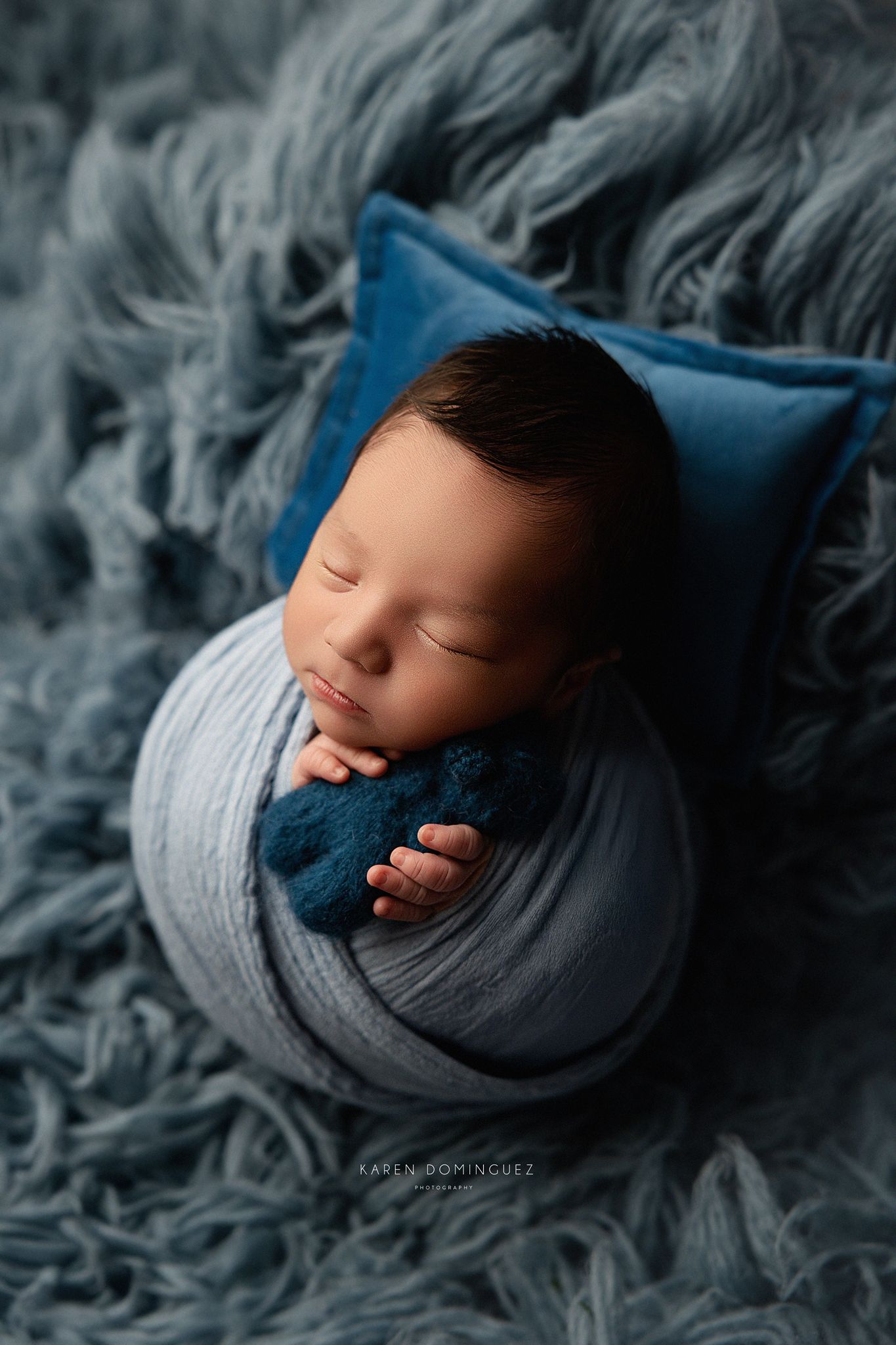 A newborn baby sleeps peacefully wrapped in gray and navy swaddles against a smokey gray textured background.
