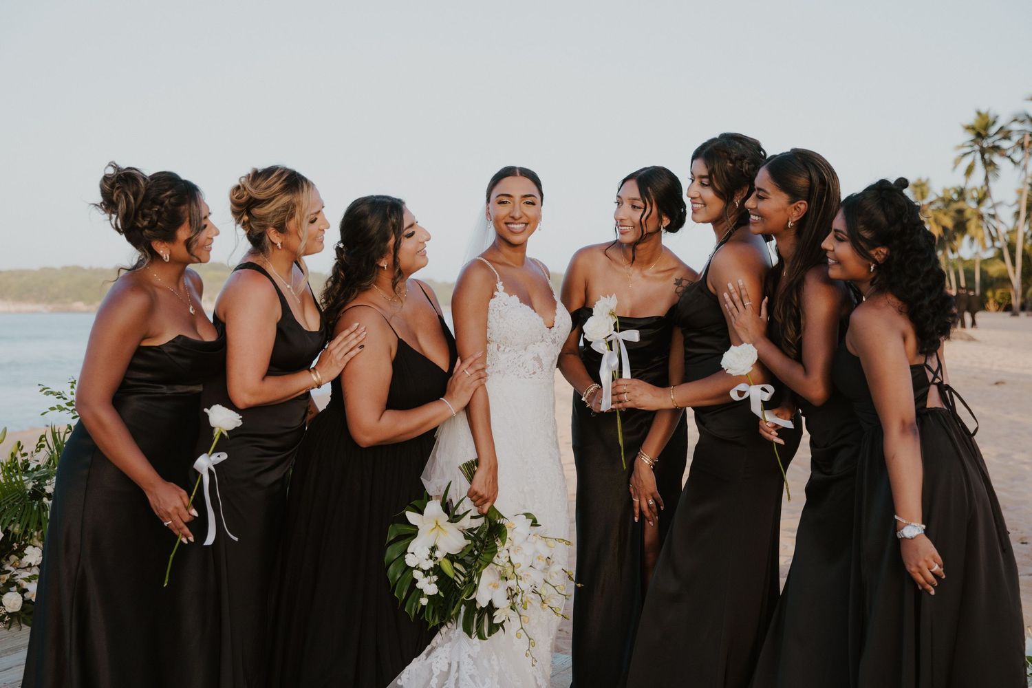 Wedding party in black dresses and suits laughing together on beach.