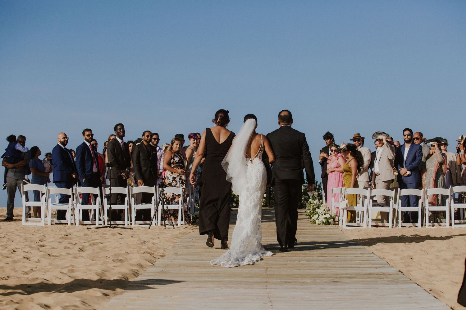 Wedding guests seated in white chairs on a beachfront wooden walkway during the ceremony.