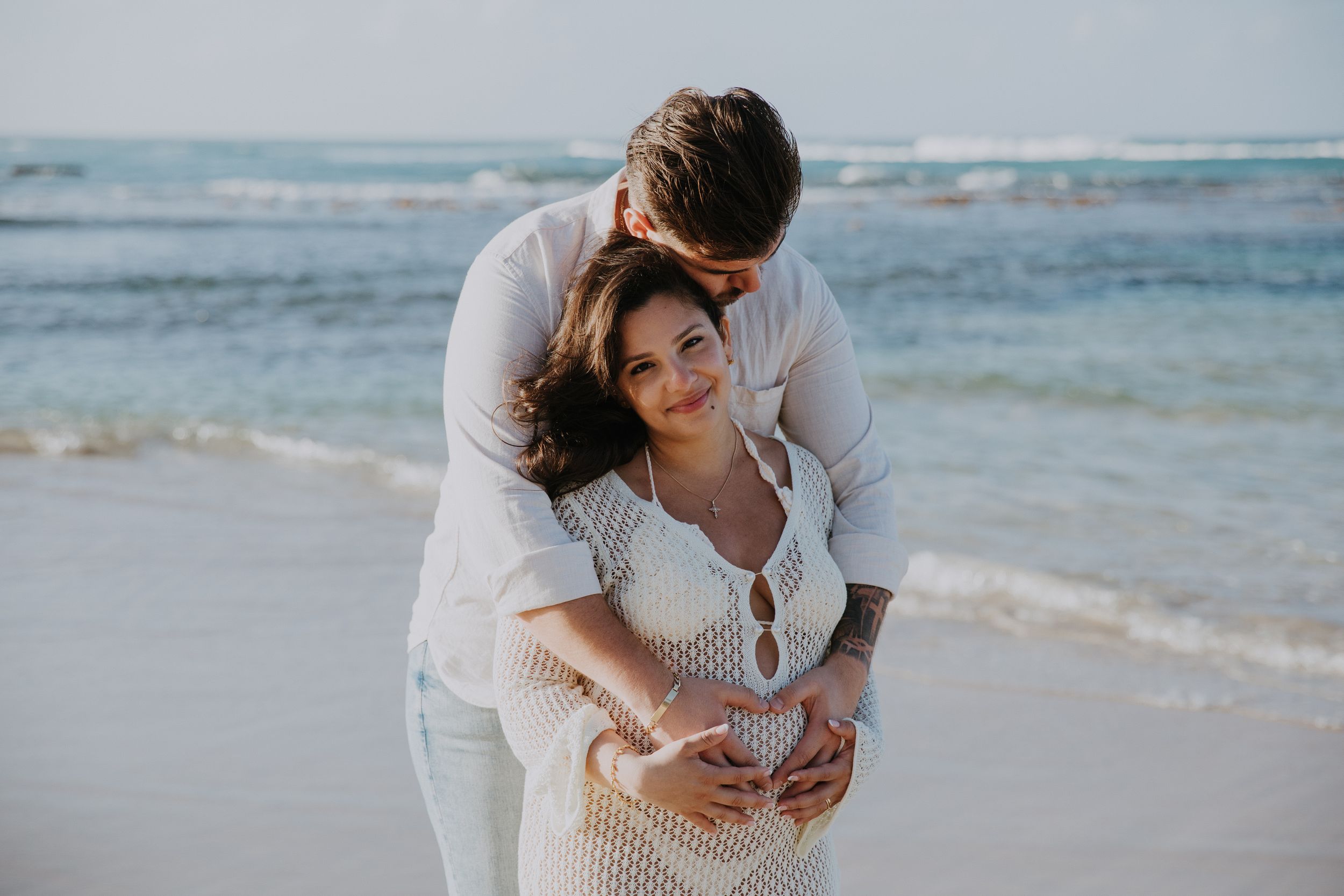 Loving embrace on beach with white lace dress and ocean waves in background.