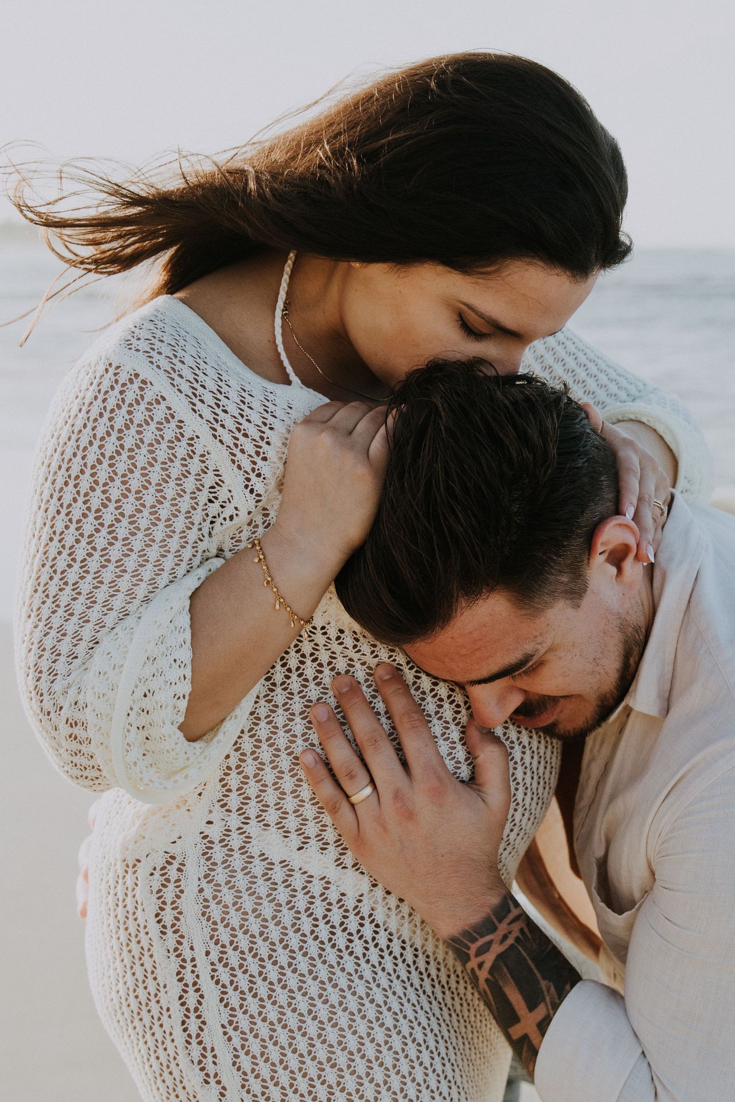 Intimate embrace captured in white lace dress against ocean backdrop.